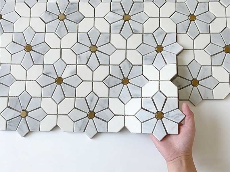 Grey and white flower-patterned stone mosaic tile with brass accents, held in hand for product display, ideal for backsplash decor.png Grey and white flower-patterned stone mosaic tile with brass accents, held in hand for product display, ideal for backsplash decor.png